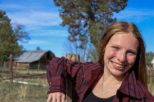 Smiling teenage girl in red plaid shirt leaning on wooden fence post with rustic barn and blue sky behind her.