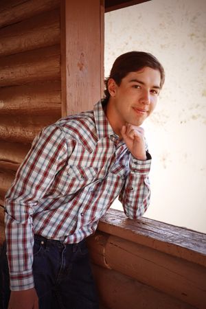 Young man in plaid shirt leaning against rustic log cabin wall with chin resting on hand.