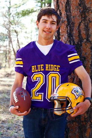 Smiling teen boy in Blue Ridge number 20 purple football jersey holding ball and yellow helmet by tree.