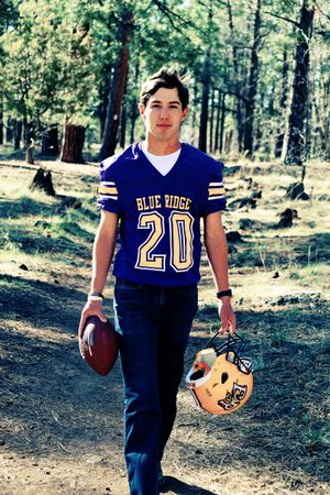 Young football player in Blue Ridge #20 jersey holding helmet and ball in forest.