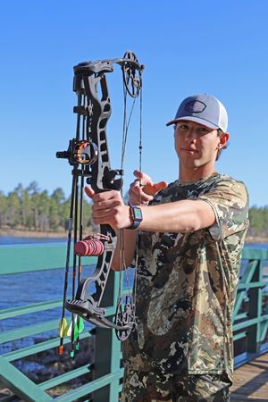 Young man in camouflage shirt holding compound bow on bridge with blue sky and lake behind him.