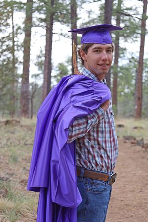 Smiling young male graduate in purple cap and gown over plaid shirt posing in pine forest.