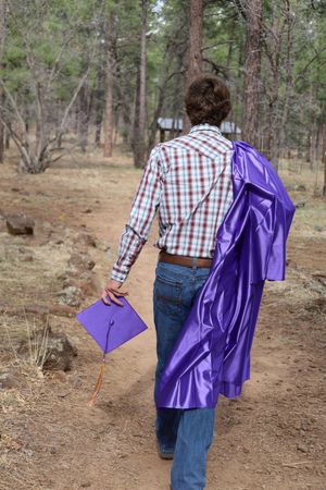 Graduate walking forest path carrying purple cap and gown in plaid shirt.