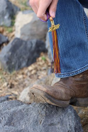 Close-up of hand holding graduation tassel beside cowboy boot on rock.