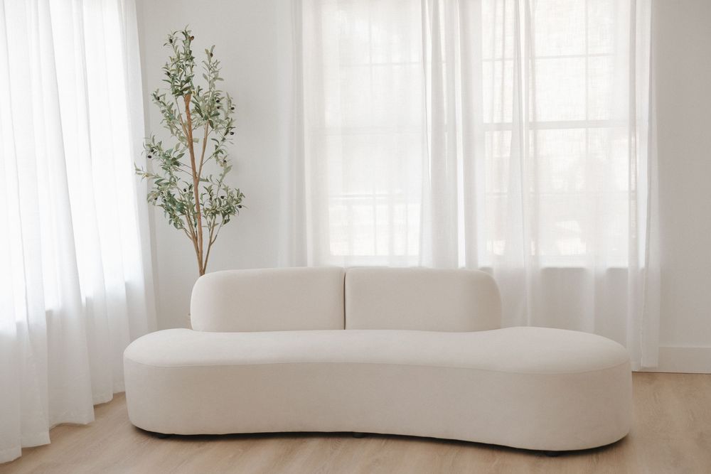 Modern white curved sofa in a bright minimalist living room with sheer curtains and a small olive tree.