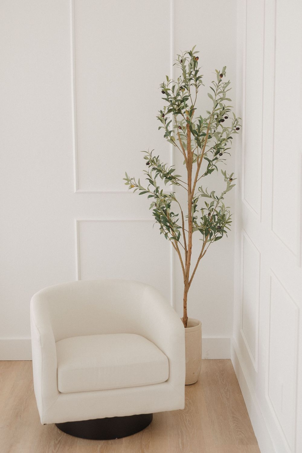 White boucle swivel accent chair beside a tall olive tree in a neutral pot against a white paneled wall.