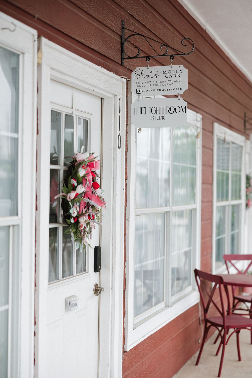 Red building exterior with white door, floral wreath, and hanging signs for Shots by Molly Carr and The Lightroom Studio.