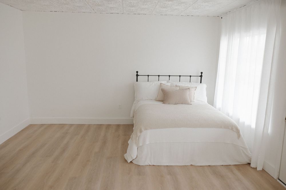 Minimalist white bedroom with iron bed frame, white bedding, light wood floors, and sheer curtains.