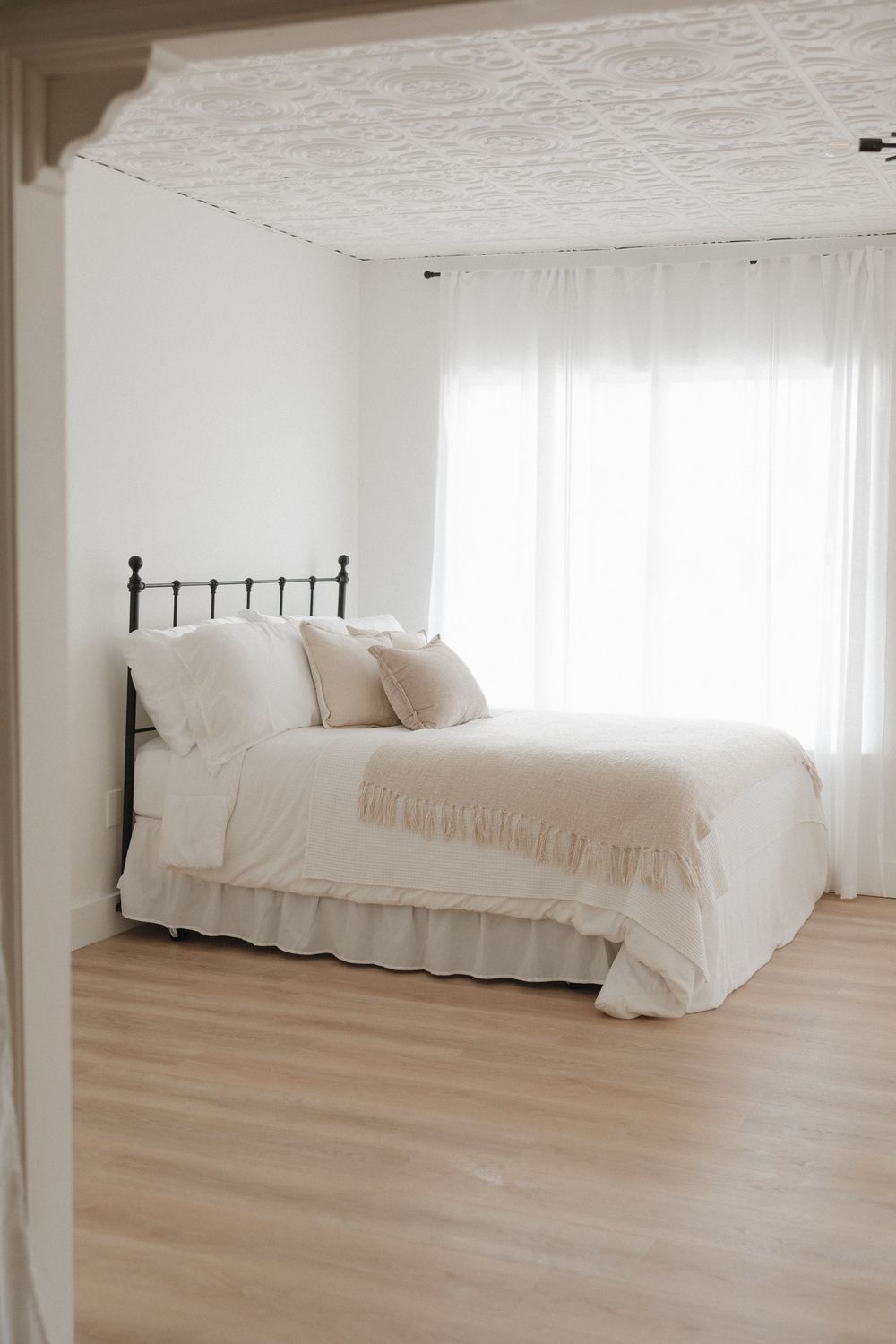 Minimalist white bedroom with iron bed frame, neutral linen bedding, fringe throw, sheer curtains, and light hardwood floors.