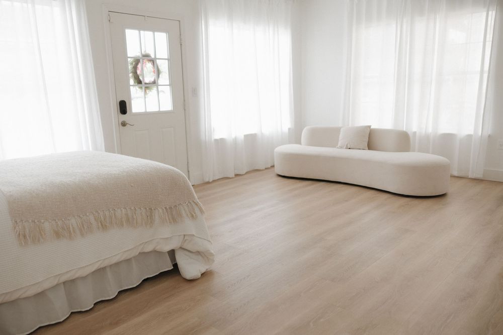 Bright white minimalist bedroom with hardwood floors, curved sofa, sheer curtains, and wreath-adorned door.
