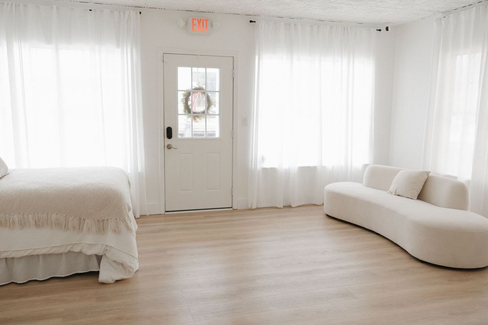 Airy white room featuring a curved sofa, bed with ruffled skirt, sheer curtains, and decorative wreath on door.