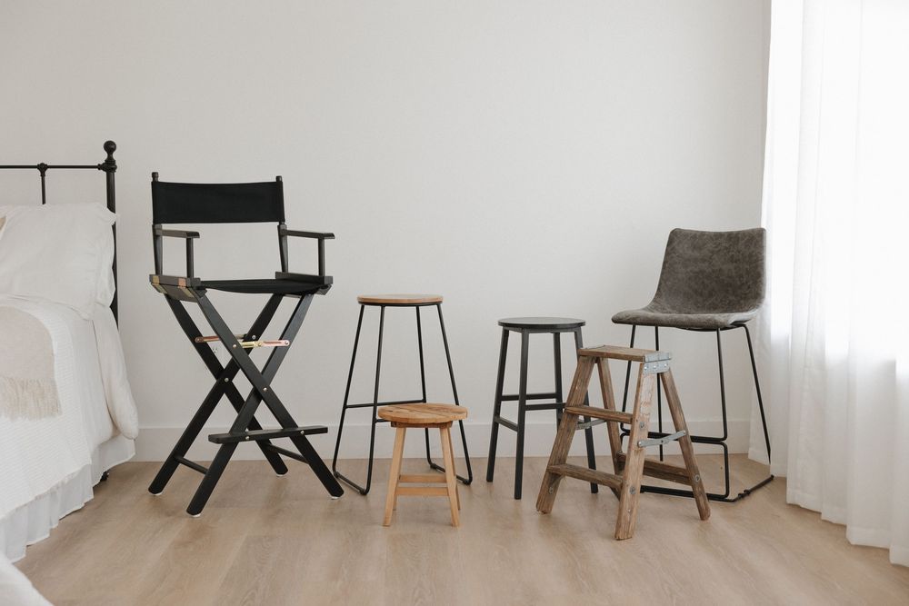 Four modern bar stools and chairs arranged against a white wall on light wood flooring in a minimalist bedroom setting.