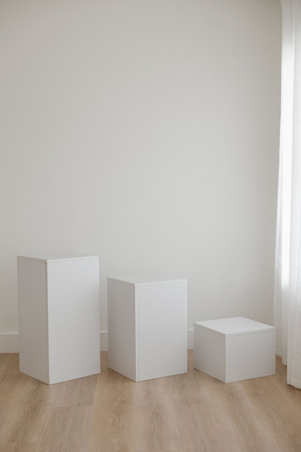 Three white rectangular pedestals of varying heights arranged on light wood flooring against a white wall.