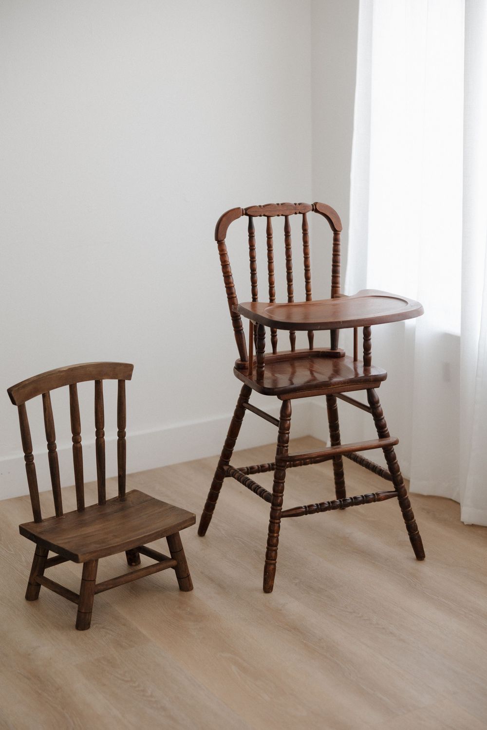 Antique dark wood spindle high chair with tray beside matching small child's chair on light hardwood floor.