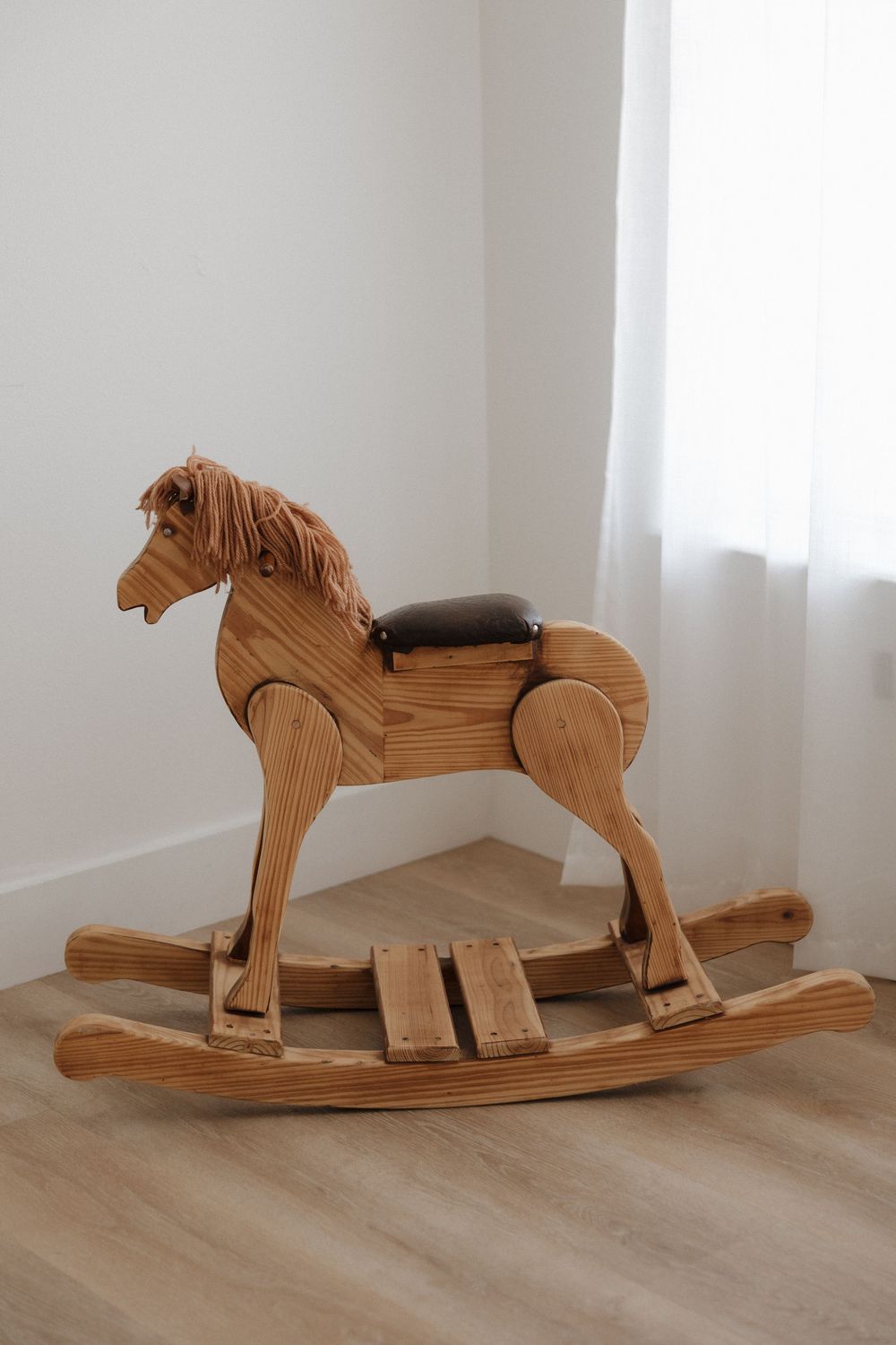 Handcrafted wooden rocking horse with brown mane and leather saddle on light hardwood floor in white room.