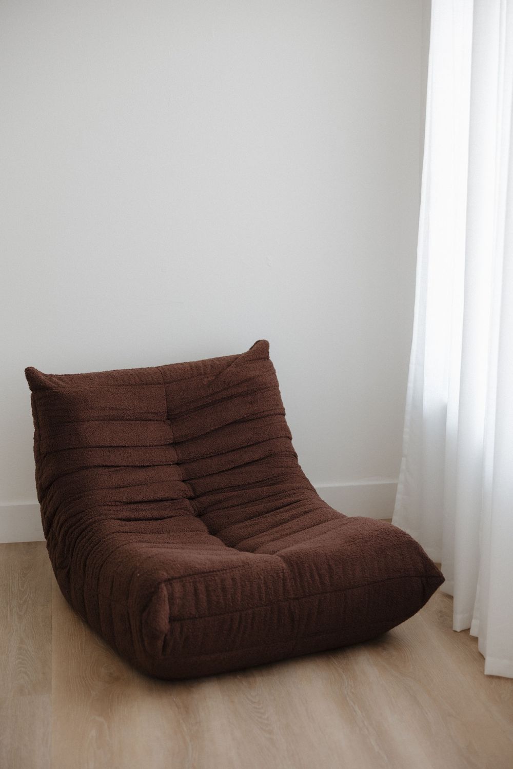 Brown ribbed foam lounge chair in minimalist white room with sheer curtains and light wood floor.