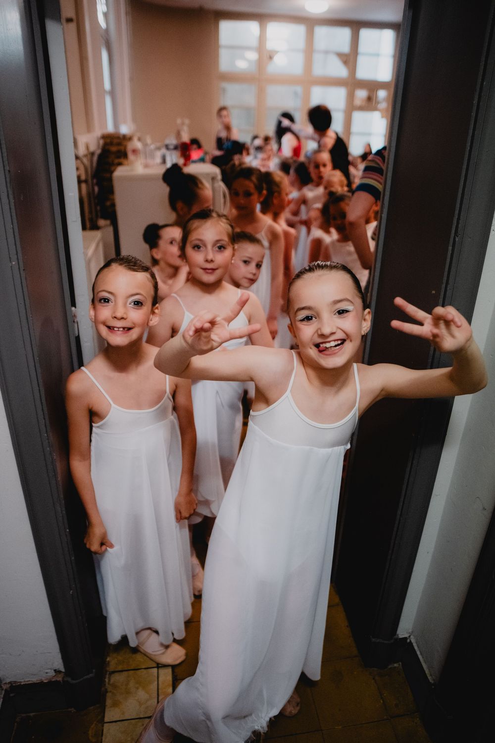 Young ballet students in white dresses gather excitedly by a doorway before their dance performance.