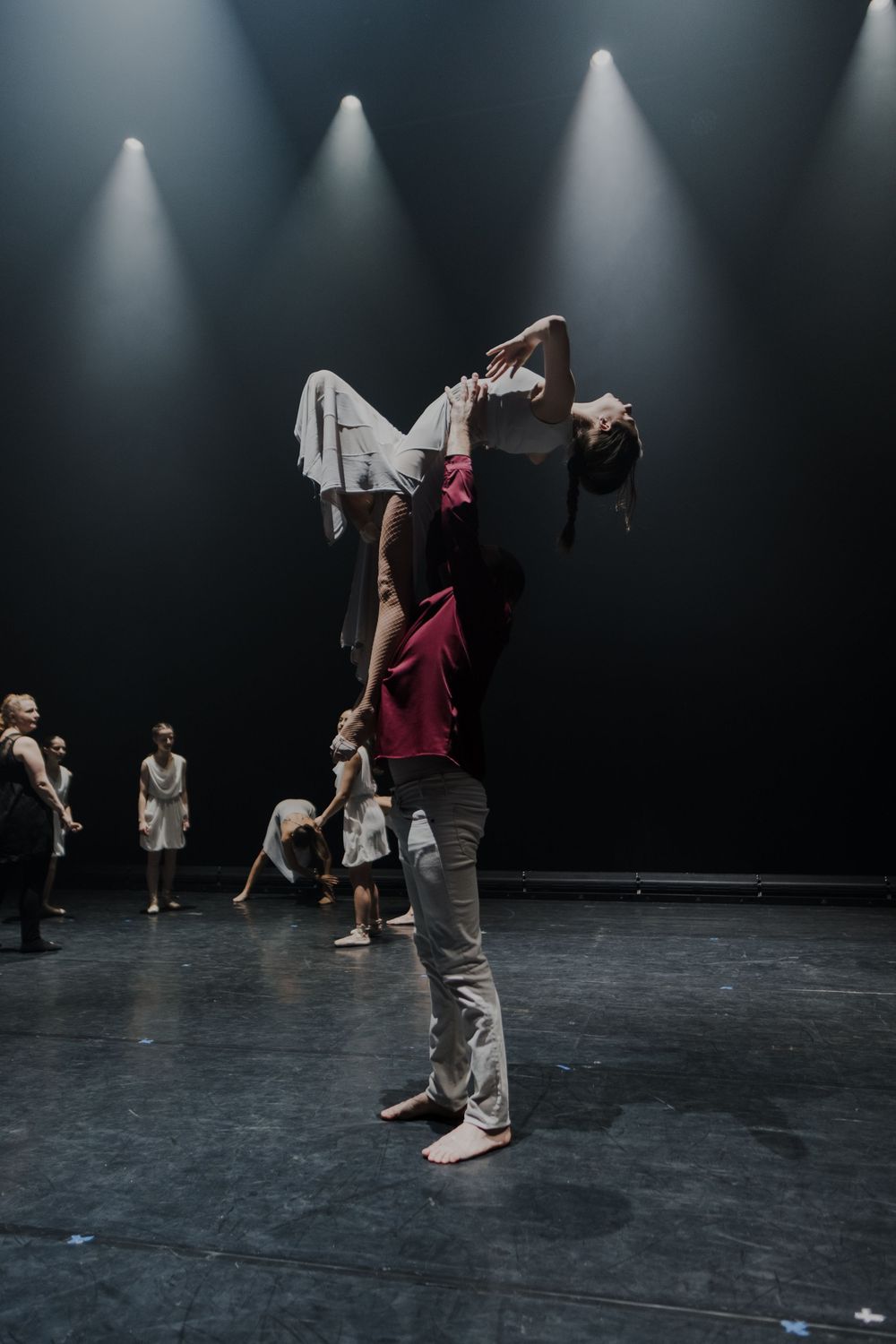 Two dancers perform a dramatic lift on a dark stage under spotlights during a contemporary dance rehearsal.