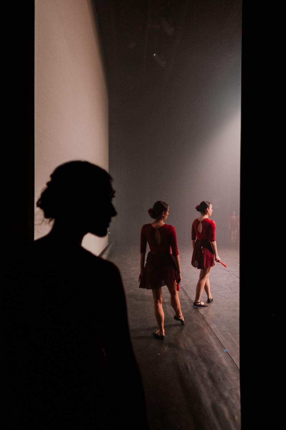 Silhouetted figures in red dresses walk across a dimly lit stage while another person watches from the shadows.
