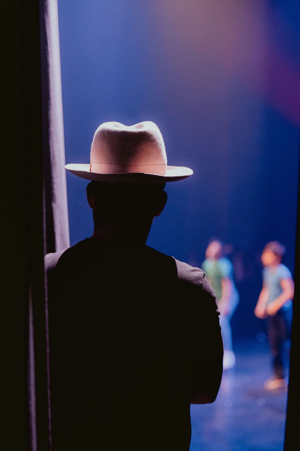 Silhouetted figure in a fedora hat stands in a doorway watching people on a blue-lit stage.