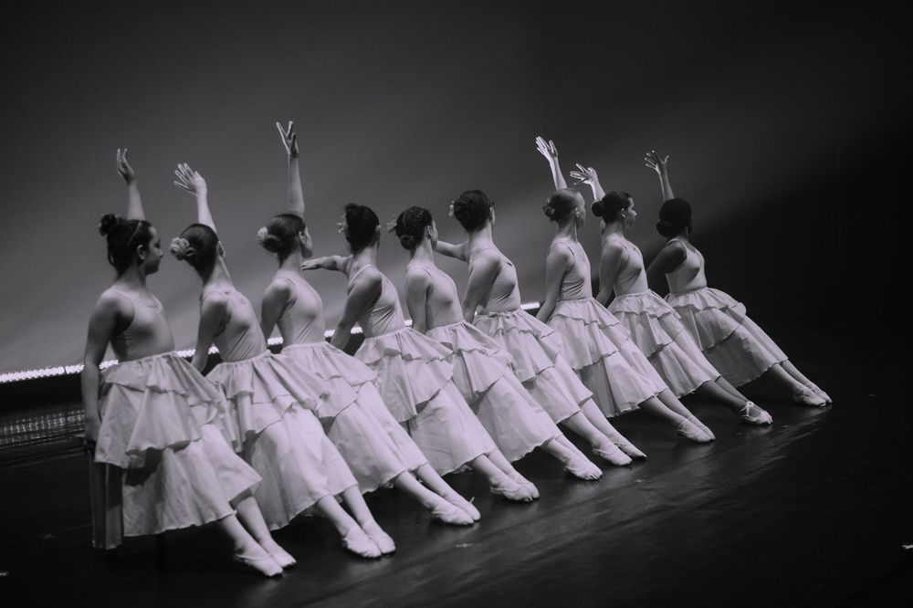 Classical ballet dancers in white tutus performing a synchronized diagonal line formation in dramatic lighting.