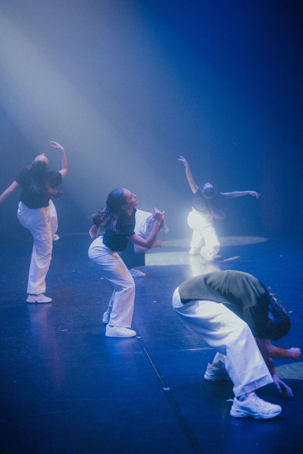 Dancers in white outfits perform synchronized movements on stage with dramatic blue lighting and fog effects.