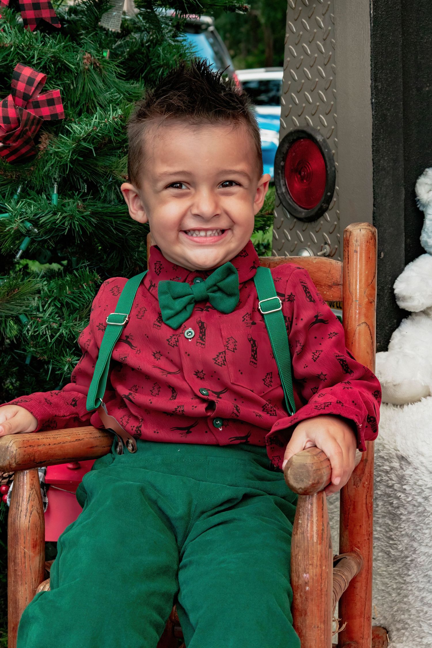 Young child in festive red and green holiday outfit sitting on wooden chair with Christmas decorations.