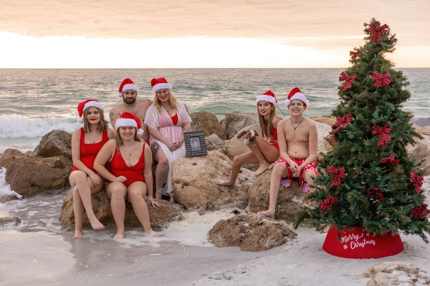 Group wearing Santa hats and swimwear posing on beach rocks next to decorated Christmas tree.