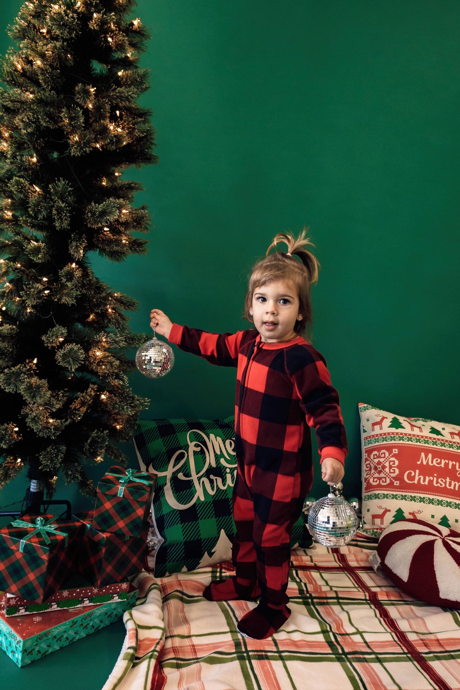 Small child in red buffalo plaid pajamas decorating Christmas tree with ornaments on green background.