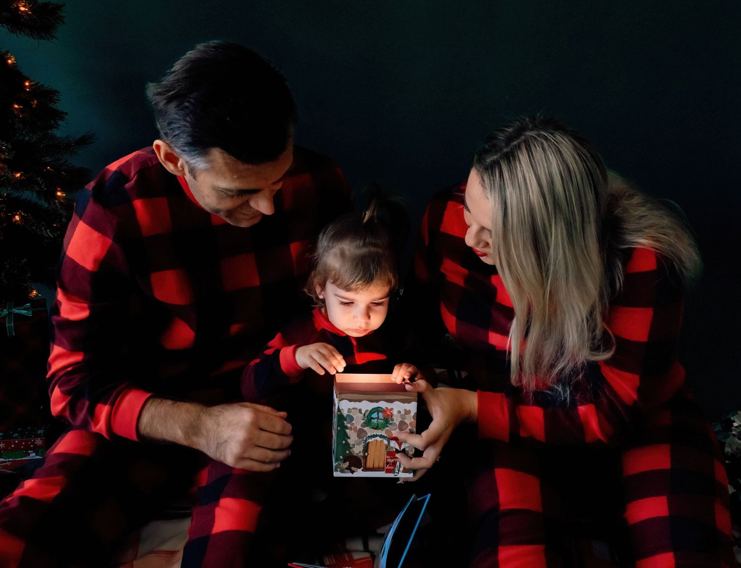 Family in matching buffalo plaid pajamas looking at Christmas gift box together.