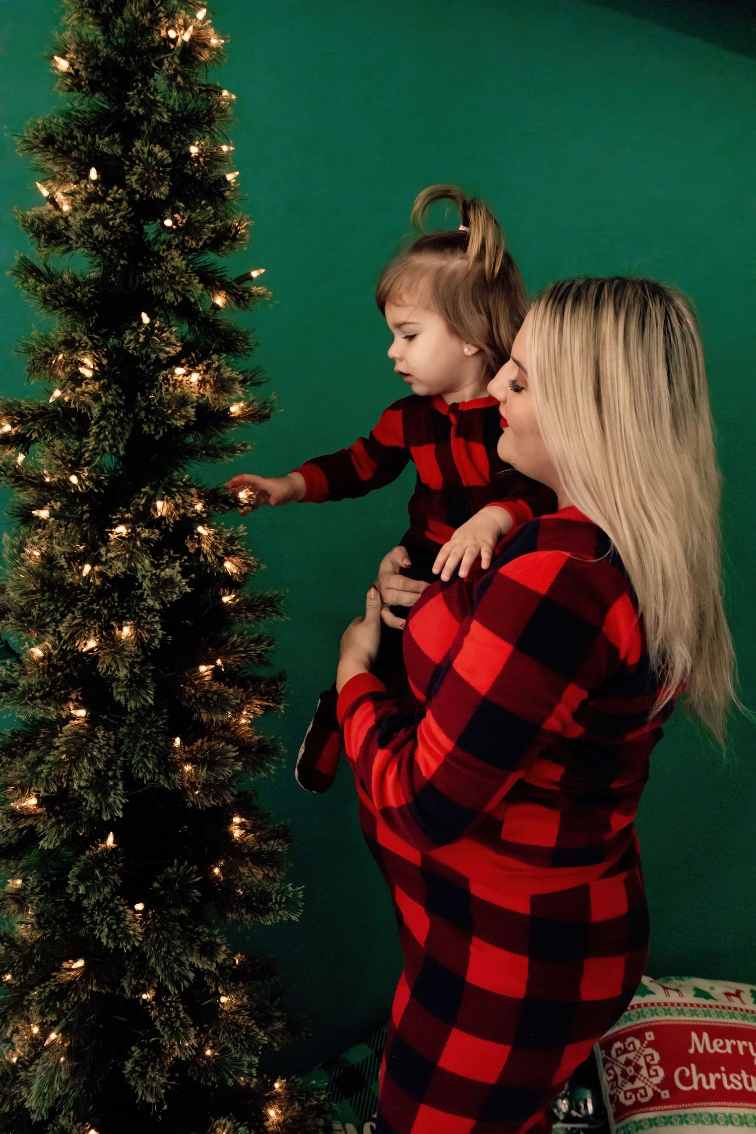 Mother and child in matching buffalo plaid outfits decorating Christmas tree against green backdrop.
