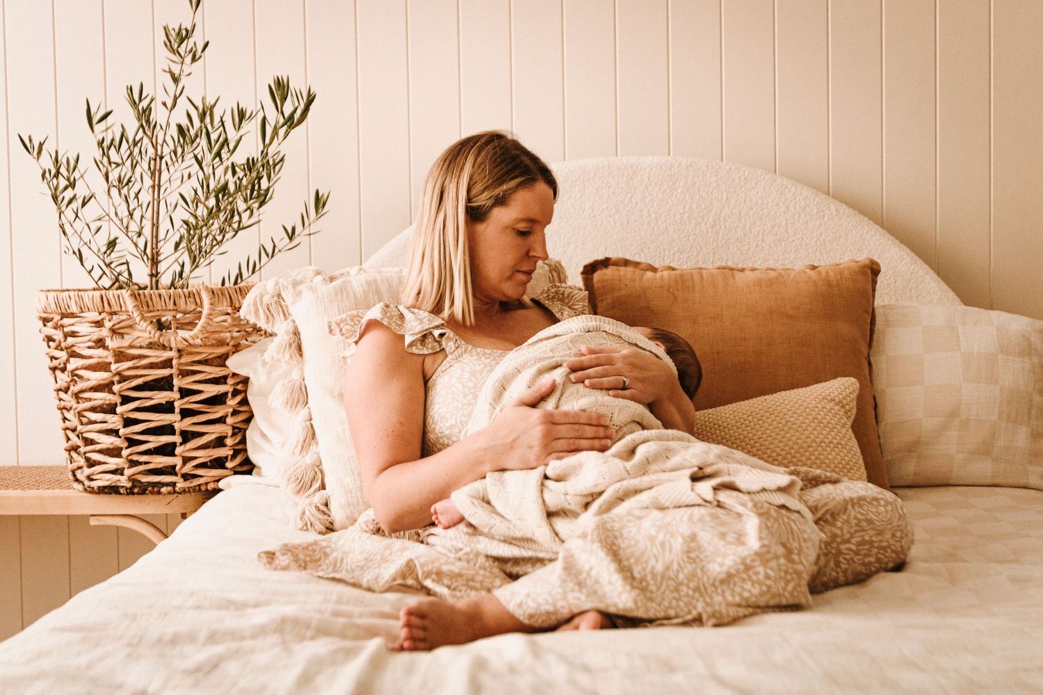 A cozy bedroom setting in warm tones features someone cuddling with a newborn baby on a plush bed.