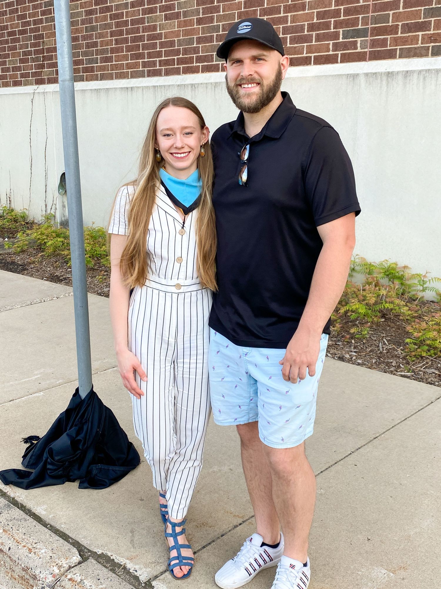 Two people stand together on a sidewalk, one wearing a striped jumpsuit and one in a black polo and blue patterned shorts.
