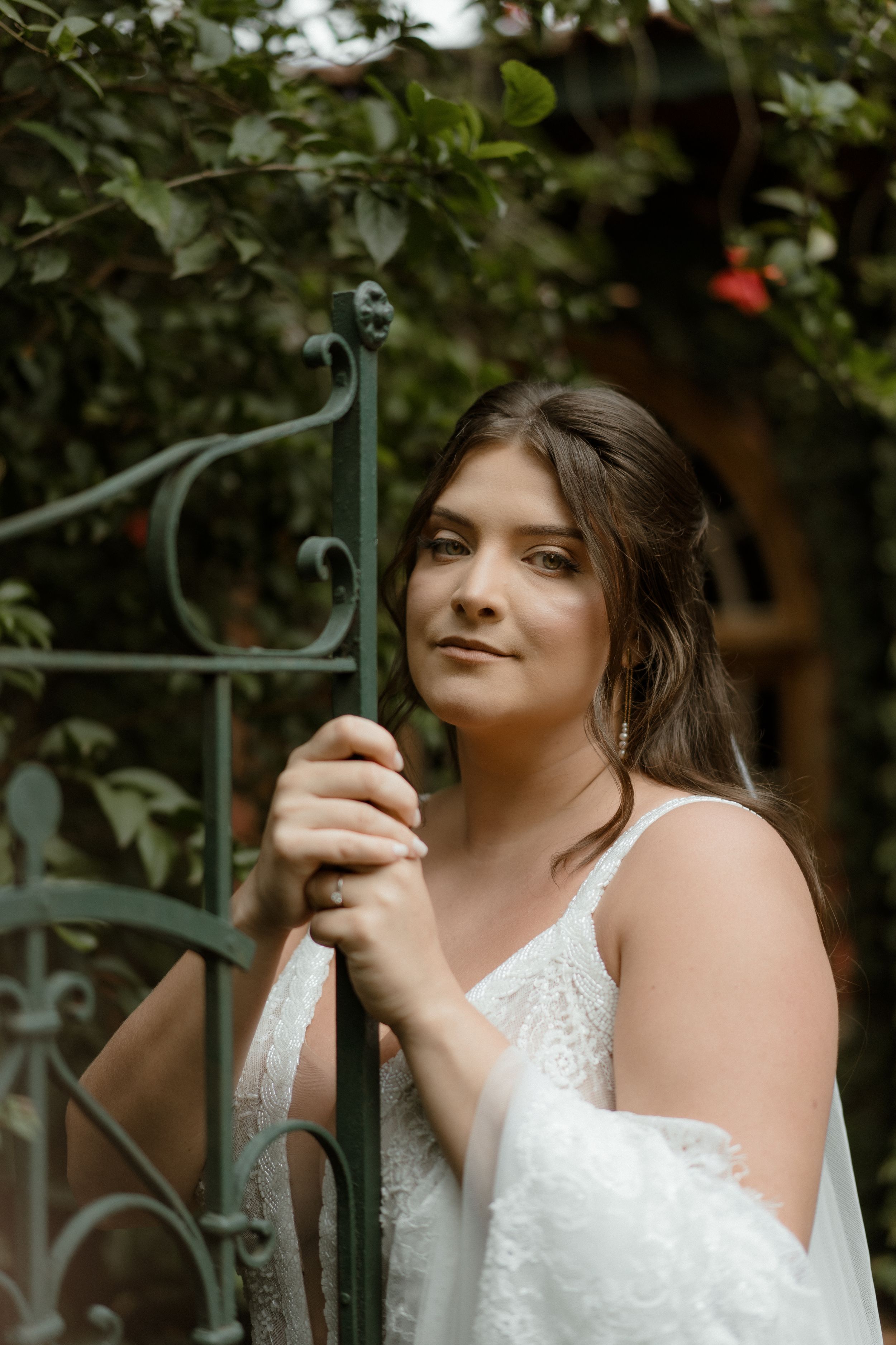 Romantic bridal portrait holding iron garden gate surrounded by greenery.