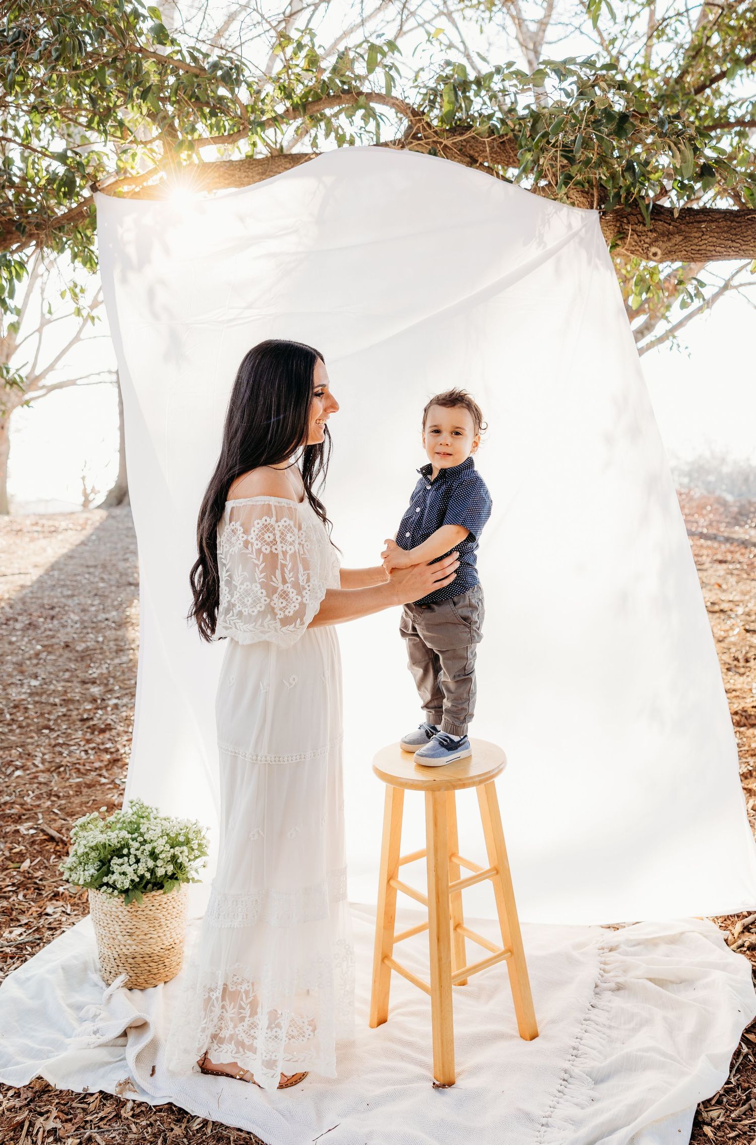 A dark-haired mother in a white lace dress holds her toddler son on a stool under a white backdrop outdoors.
mommy and me
simple timeless