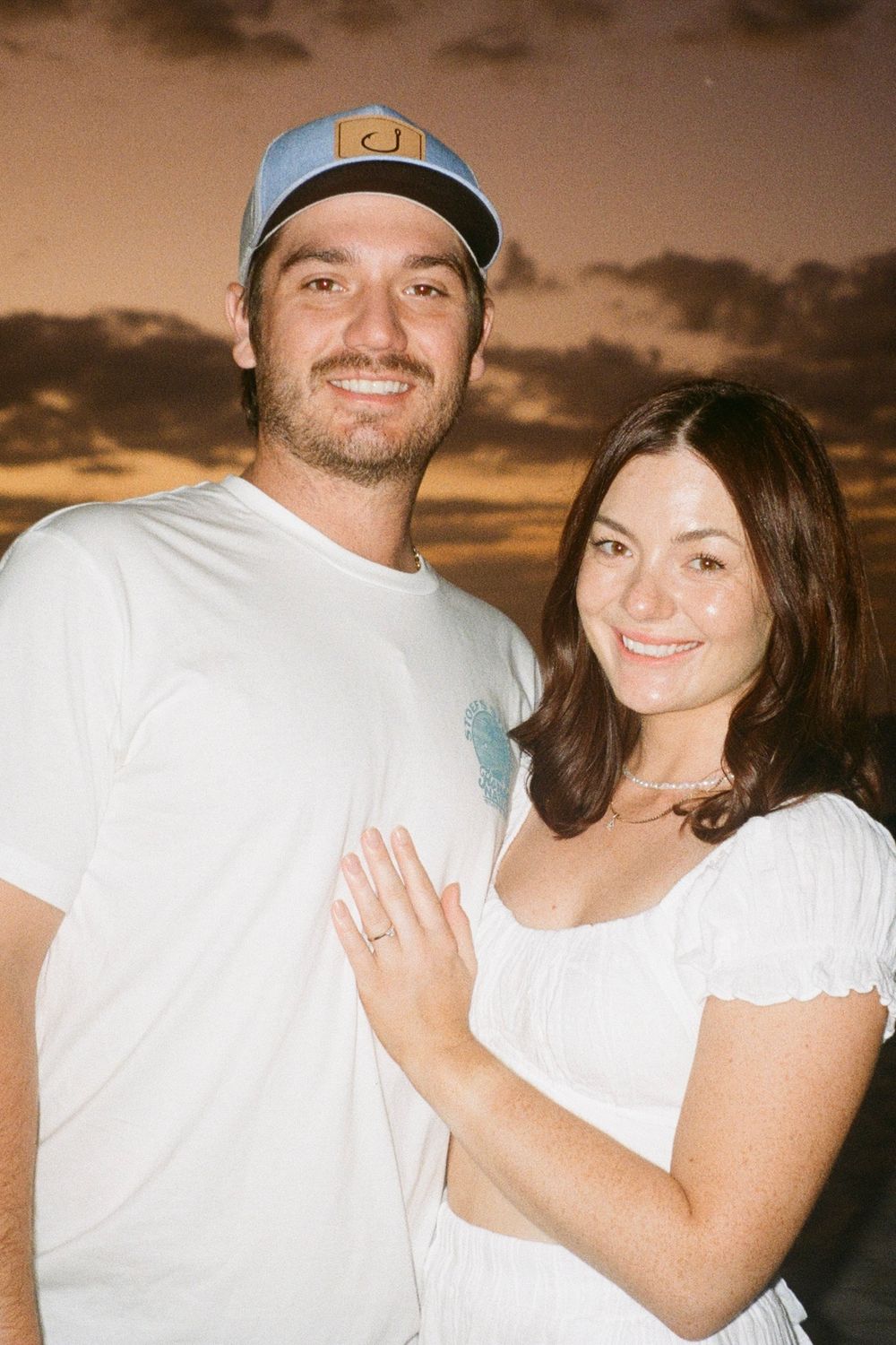 A couple wearing white clothing poses together against a dusky sunset sky.