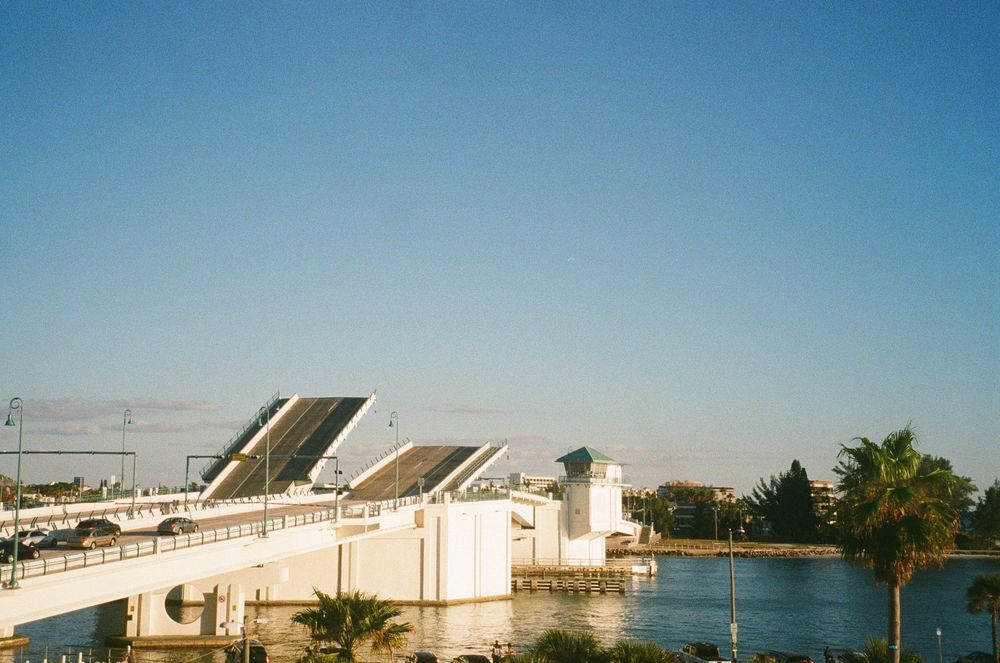 A white drawbridge spans across a waterway under clear blue skies with palm trees visible along the shoreline.