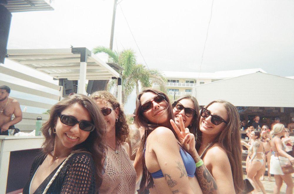 A group of friends wearing sunglasses pose together at an outdoor summer beach party or resort.