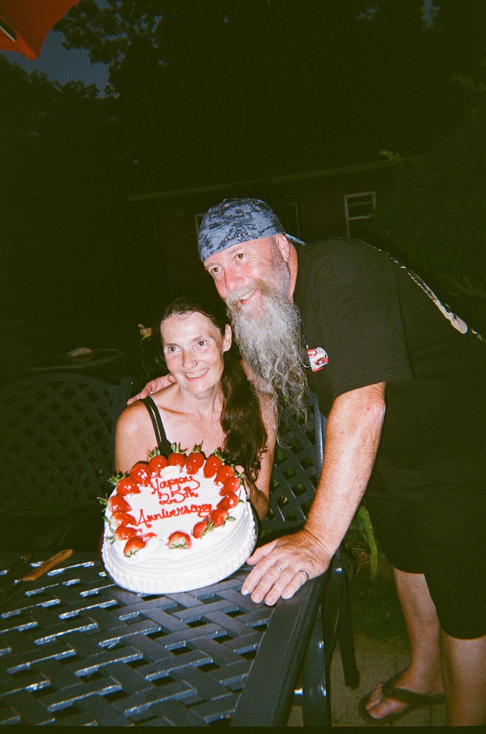 Two people celebrate with a round white and red anniversary cake on a picnic table at night.