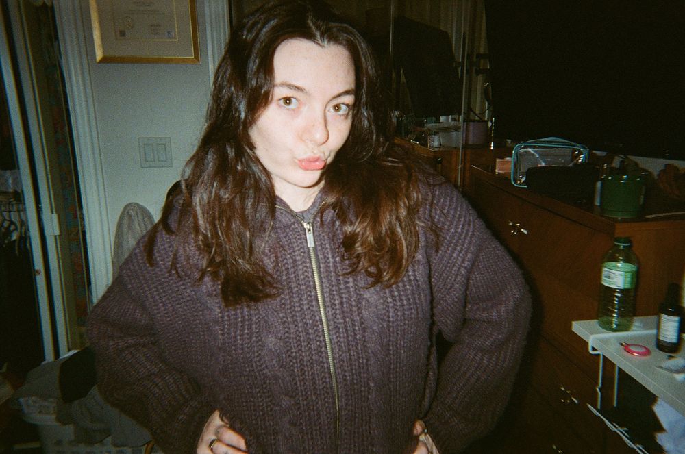 Indoor film photograph of someone in a dark brown knitted zip-up sweater in low light.