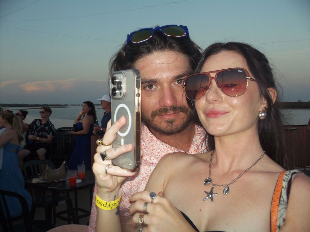 Two people take a selfie near water at sunset while holding up drinks at a waterfront venue.