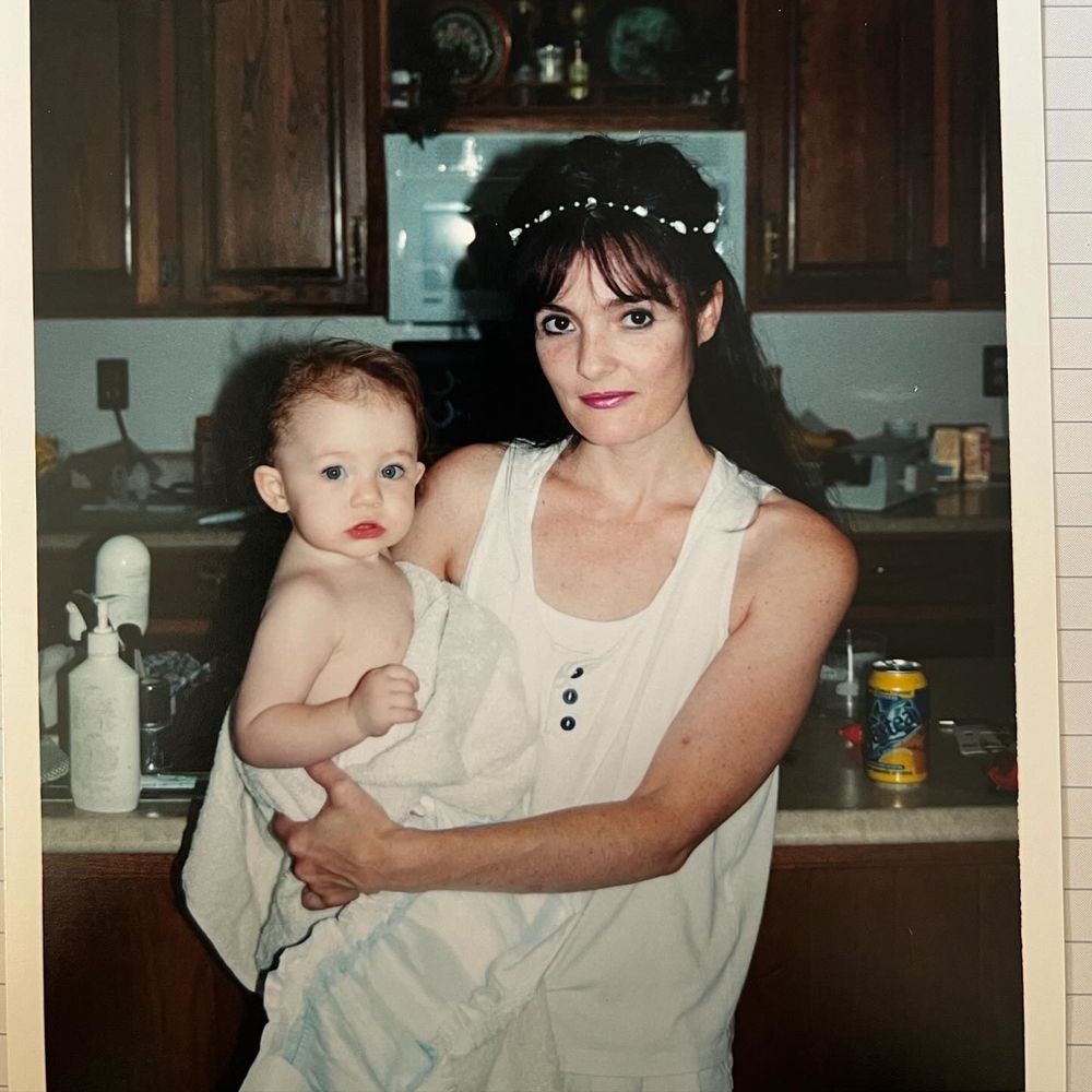 A vintage-style photo taken in a kitchen shows someone in a white dress and floral headband holding a baby.