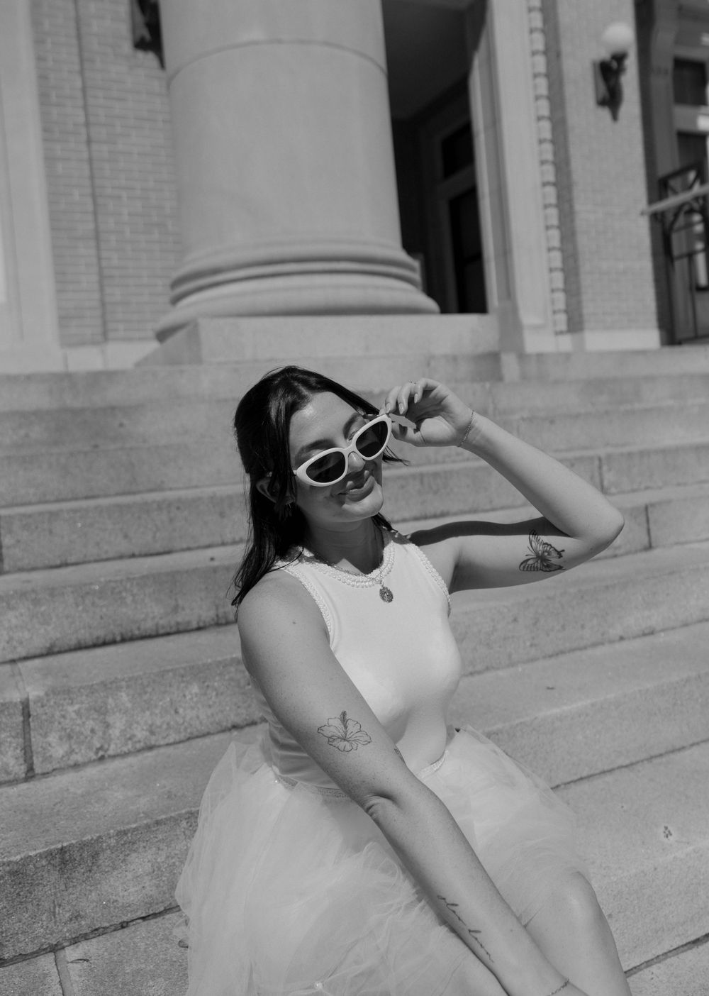 Stylish woman in white dress and retro sunglasses poses on stone steps near classical building column in black and white.