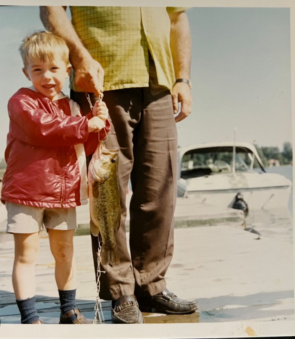 Child in red jacket and shorts poses with a fish caught on a pier with a boat visible in the background.