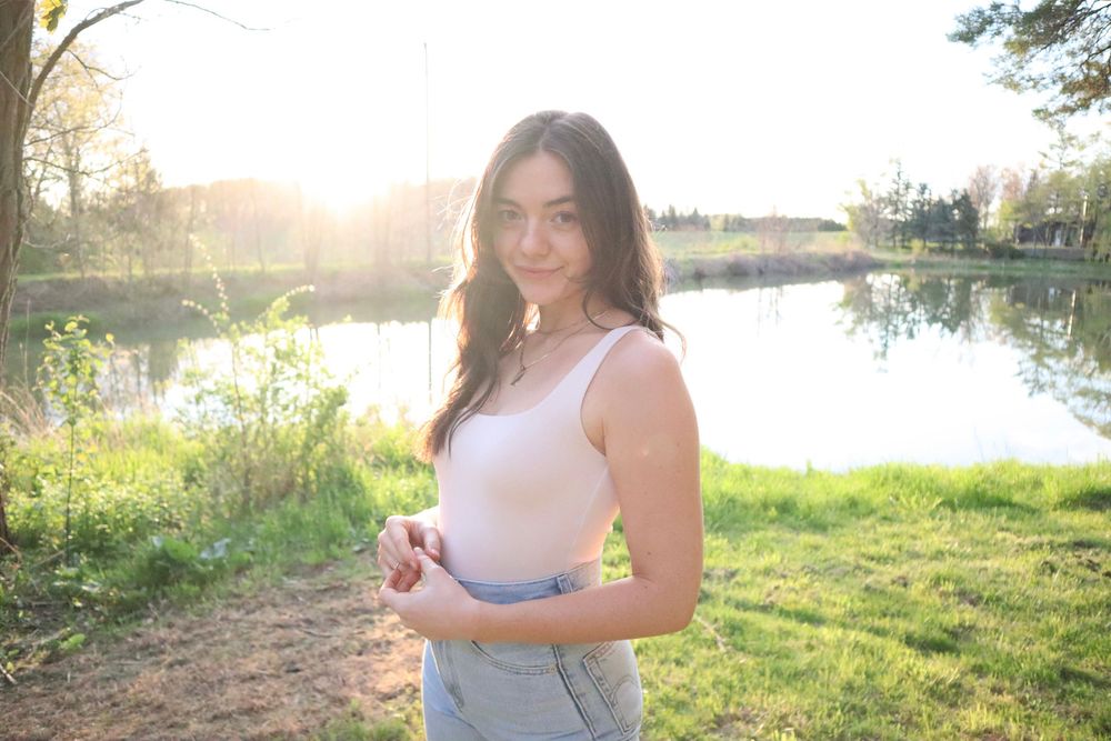 Person in casual attire stands by a serene lake at sunset with golden sunlight filtering through trees in background.