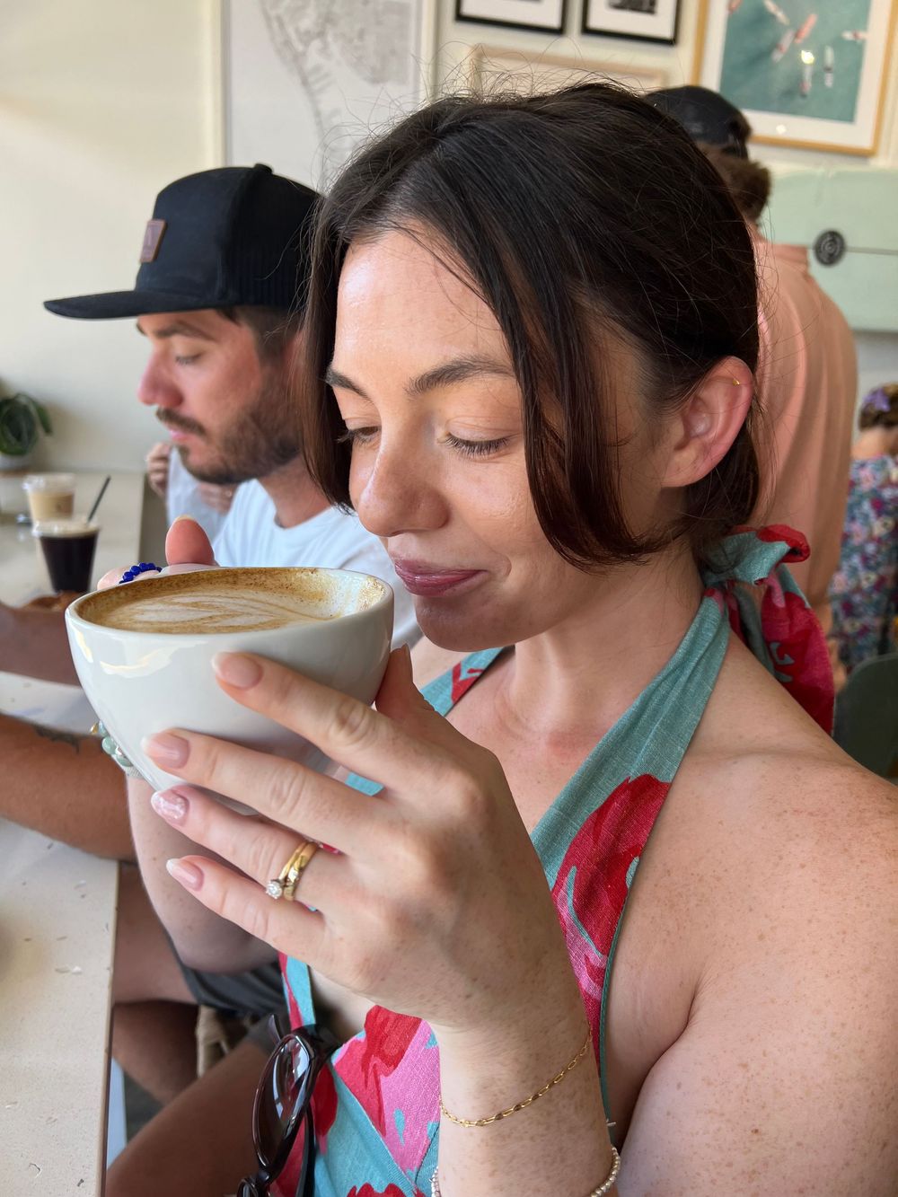 A person in a floral dress holding and enjoying a frothy latte at a cafe.