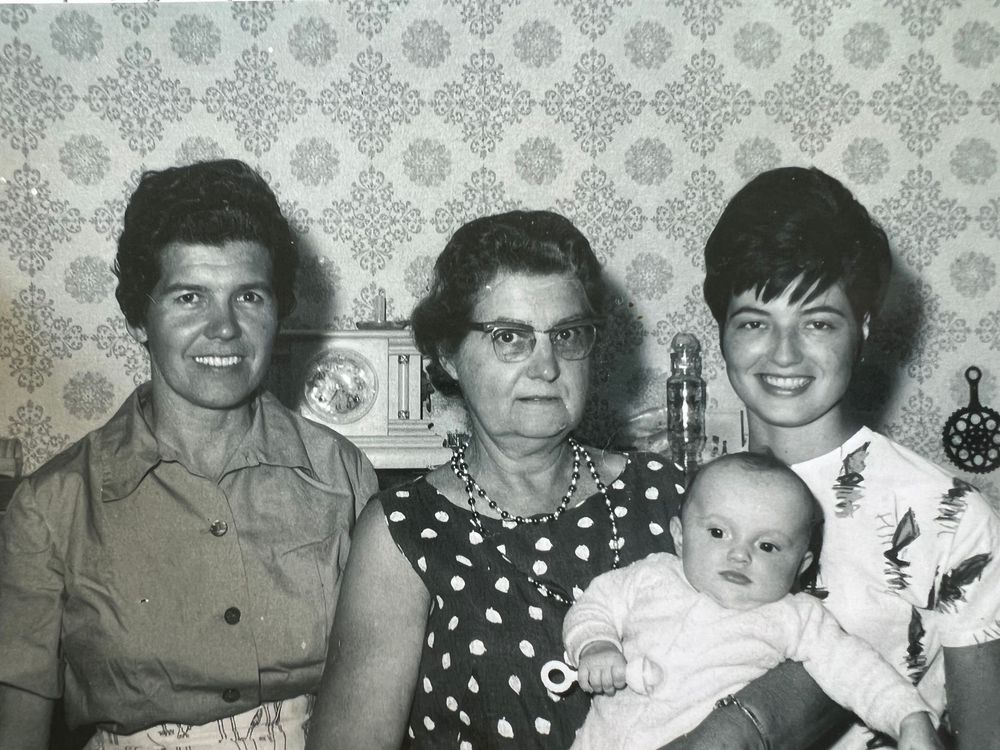 Black and white vintage family photo showing four generations together in front of decorative wallpaper.