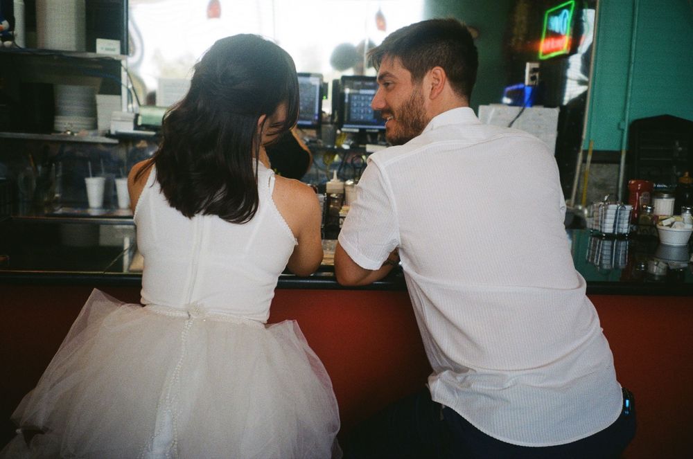 Couple in white outfits sitting together at retro diner counter with vintage film photography aesthetic.
