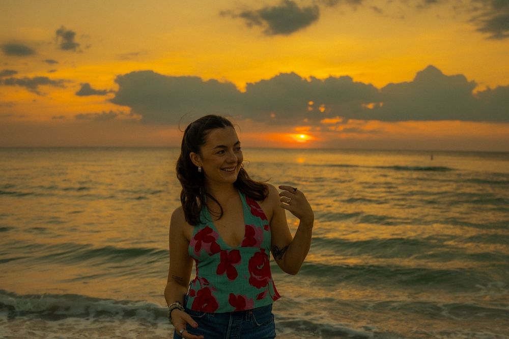 A silhouetted figure in floral attire stands on a beach at sunset with golden clouds and waves in the background.