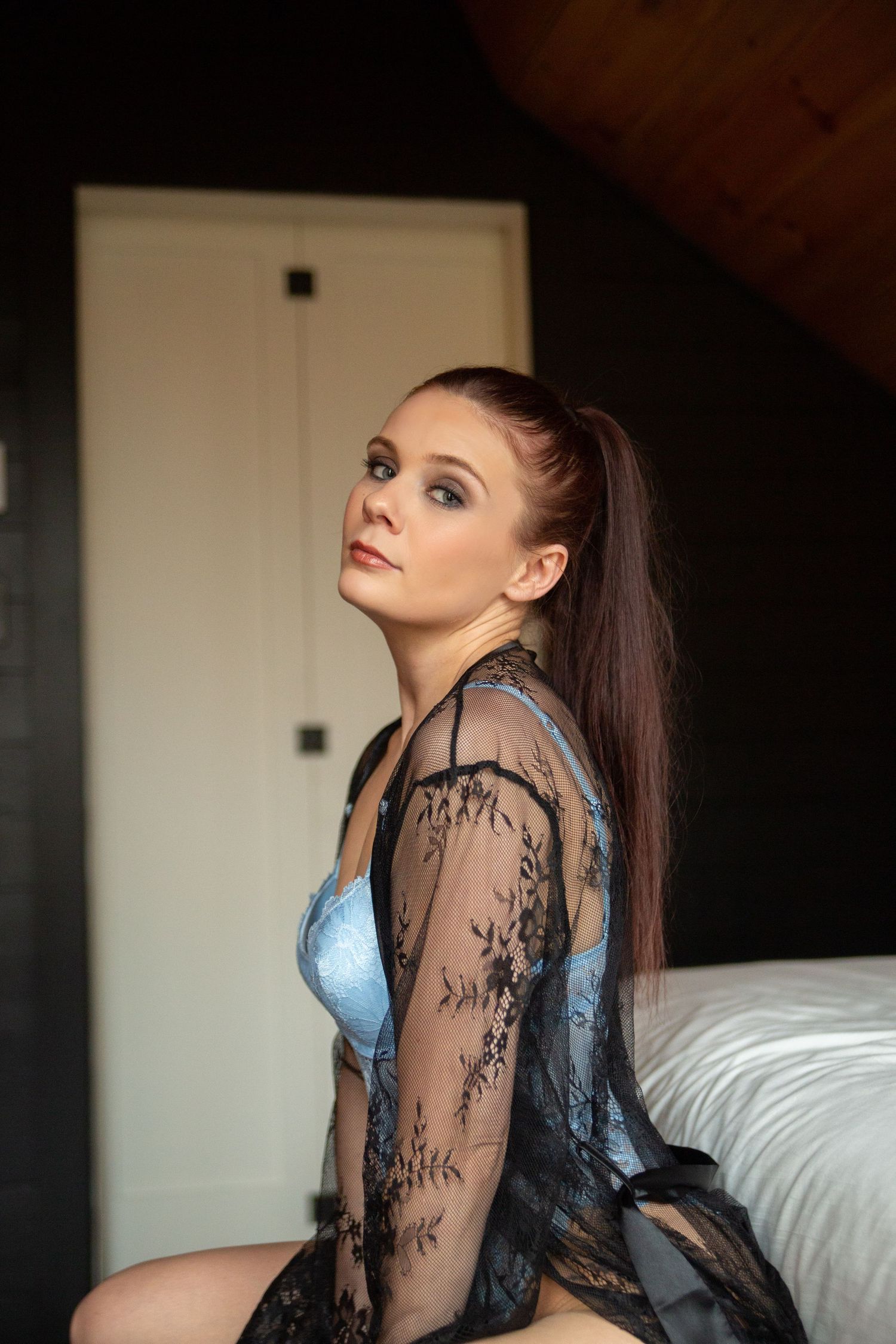 A woman in a black lace robe and blue lingerie poses seductively on a bed.